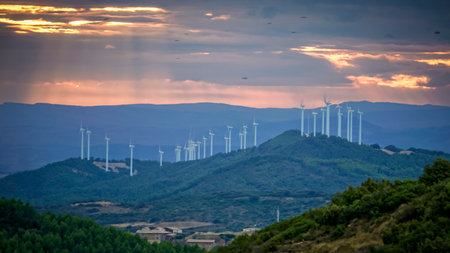 Landscape of a mountain range at sunset with the sun peeking through the clouds and some wind turbines in the distanceの写真素材