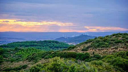 Landscape of a mountain range at sunset with the sun peeking through the clouds and some wind turbines in the distanceの写真素材