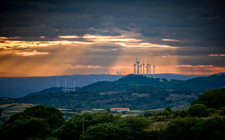 Landscape of a mountain range at sunset with the sun peeking through the clouds and some wind turbines in the distanceの写真素材