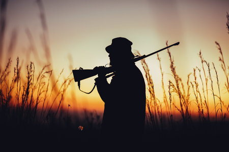 Hunter holding shotgun on shoulder and observing land in sunrise. High grass is sourrounding him