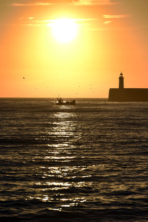 Fishing boat near lighthouse and breakwater at sunsetの写真素材