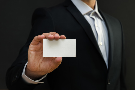 Man wearing a suit holding white business card on black wall background. Mock up, space for textの写真素材