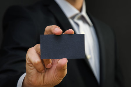 Man wearing a suit holding white business card on black wall background. Mock up, space for text