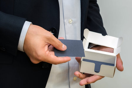 Man wearing a suit holding white business card on black wall background. Mock up, space for text