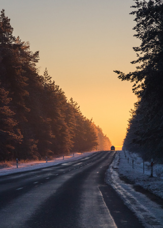Silhouette of a car on asphalt road in snowy winter sunset light. Scenic spruce trees with cold snow on branches by the roadの写真素材