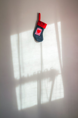 Red Christmas sock with a heart hanging on white wall with shadow of a window