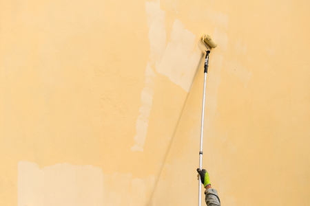 Painter paints building exterior wall with a roller. Roller with long stick manually painting building with yellow paintの写真素材