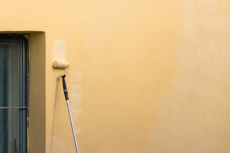 Painter paints building exterior wall with a roller. Roller with long stick manually painting building with yellow paintの写真素材