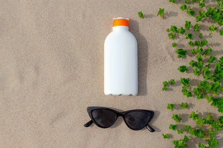 Sunscreen protection lotion and sunglasses in the sand on a sunny day at the beach. White sunblock...