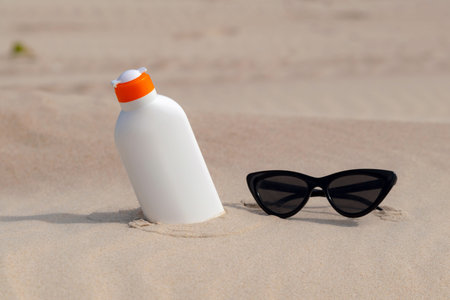 Sunscreen protection lotion and sunglasses in the sand on a sunny day at the beach. White sunblock...
