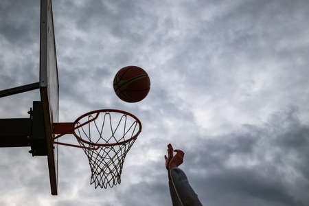 Dark silhouette of street basketball ball falling into the hoop on a cloudy day. Close up of a ball above the hoop net. Concept of success, scoring points and winning. Urban youth game.の写真素材