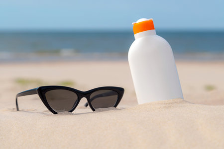 Sunscreen protection lotion and sunglasses in the sand on a sunny day at the beach. White sunblock...