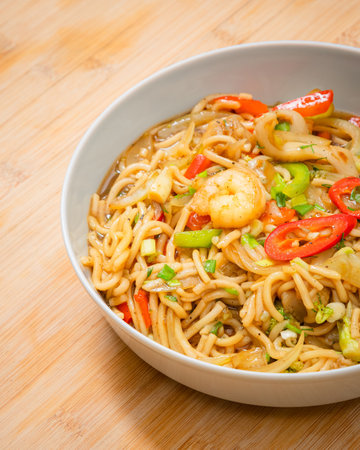 Golden coloured spicy sea food noodles with shrimp, broccoli, chilli pepper in a plate on wooden tableの写真素材