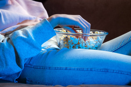 Close up o f hand waking popcorn from a bowl while watching TV. Person sitting in comfortable couch and watching home cinema in the dark.の写真素材