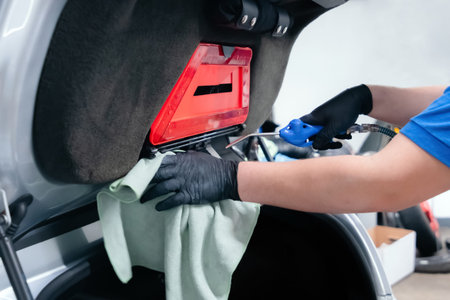 Close up of car wash worker wearing protective gloves and cleaning car trunk with compressed air and...