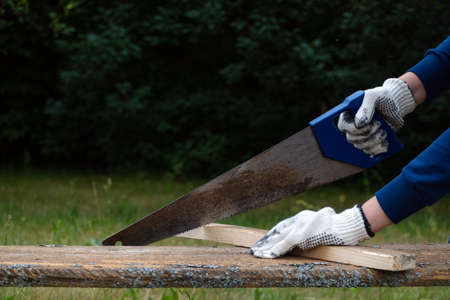 Close up of carpenter wearing gloves and hand sawing wooden plank with old blunt saw in the garden.の写真素材