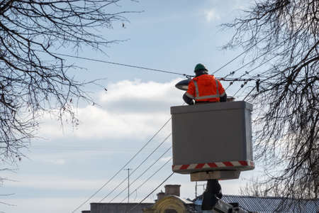 Electrician in a lift platform wearing protective work clothes changing public street lights on a sunny day.の写真素材