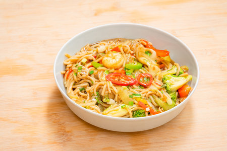 Golden coloured spicy sea food noodles with shrimp, broccoli, chilli pepper in a plate on wooden tableの写真素材