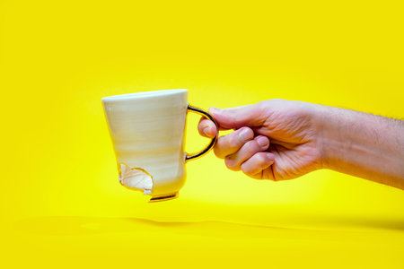Man holding broken tea cup isolated on yellow background. Close up of arm holding cracked coffee mugの写真素材