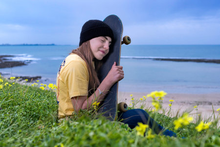 Teenager girl with skateboard sitting on a grass near the sea