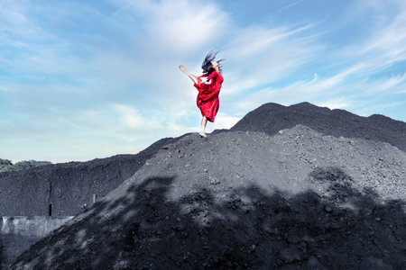 Full body of female with black flying hair in red dress raising leg while dancing on pile of coal...