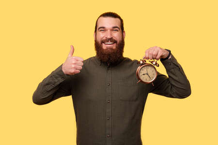 A picture of a happy man holding a clock and his thumb upの写真素材