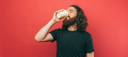 Banner size photo of bearded hispter man with long hair drinking cup of coffee to go over pink backgroundの写真素材