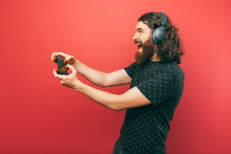 Side view photo of amazed bearded man playing games with joystick and screaming over pink backgroundの写真素材