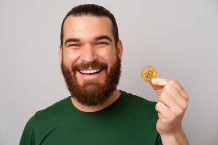 Close up photo of joyful smiling bearded man holding bitcoin over grey backgroundの写真素材