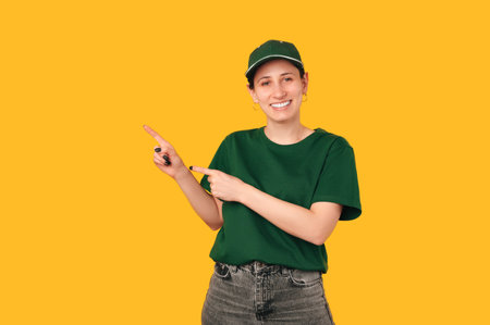 Cheerful young woman wearing delivery uniform points aside with two fingers.