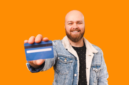 Photo of happy amazed young bearded man showing blue debit card over orange backgroundの写真素材