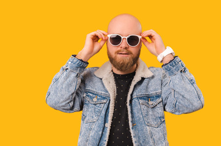 Handsome bearded bald man wearing jeans is holding cool white sunglasses.