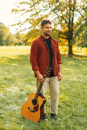 Vertical outdoors portrait of a young man holding an acoustic guitar on grass.