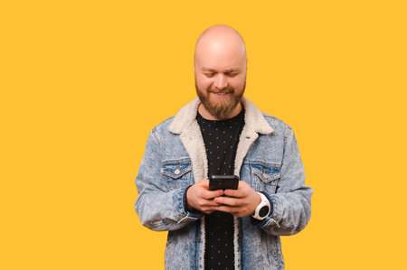 Handsome bearded bald man is texting and checking his phone in a yellow studio.