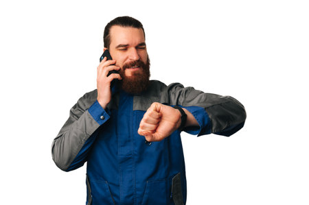 Handsome bearded handyman is talking on the phone and checking his wrist watch.の写真素材