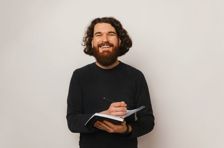 Portrait of smiling young bearded man making notes in planner over white background