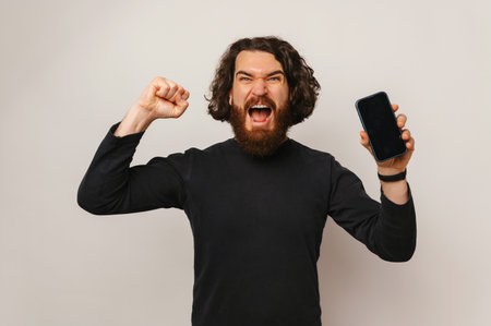 Photo of amazed young bearded hipster man celebrating victory and holding smartphone over white backgroundの写真素材