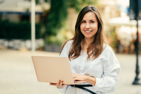Young woman holding her open laptop is standing and smiling at the camera.の写真素材