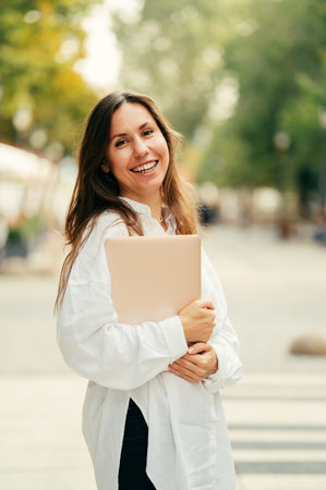 Vertical street lifestyle portrait of a young smiling woman holding her laptop.の写真素材