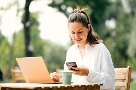 Happy young woman is working online while sitting at a cafe terrace in a park.の写真素材