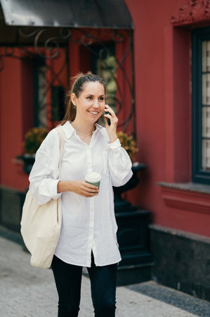 Smiling woman is talking over the phone while holding a coffee and a tote bag.の写真素材