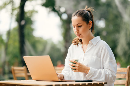 Concentrated young woman is sitting at a cafe terrace working on laptop.の写真素材