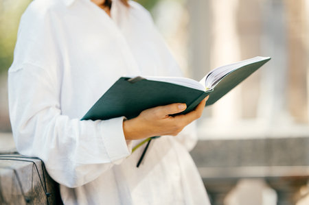 Young woman wearing white shirt is holding a green book or journal outdoors.の写真素材