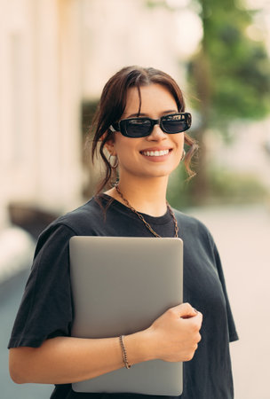 Outdoors portrait of a young smiling woman holding her laptop while walking.の写真素材