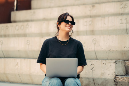 Cheerful woman works on the laptop on her knees outdoors on some stairs.の写真素材