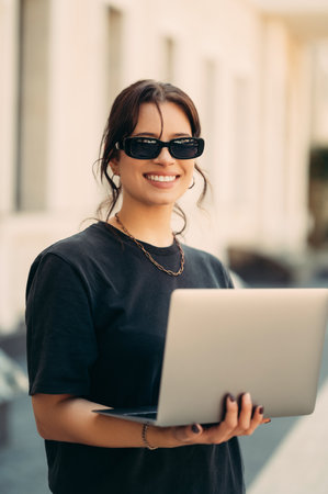 Young smiling woman is holding her laptop while standing on the street.の写真素材
