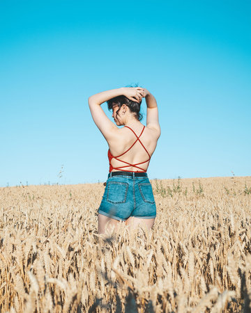 A young girl with her back turned in the middle of a wheat field on a summer day with blue sky...