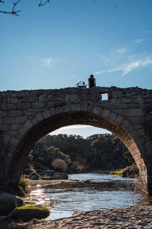 A boy stands on the arch of a Roman bridge with his toy pirate ship looking to the right on a hot summer day's sunsetの写真素材