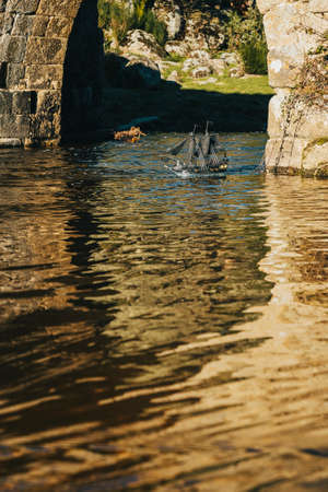 A black toy pirate ship sailing on the river under a Roman bridge on a summer day in the middle of natureの写真素材