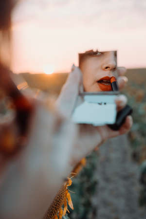 A beautiful young girl paints her lips in front of a mirror in a field of sunflowers on a summer day's sunsetの写真素材